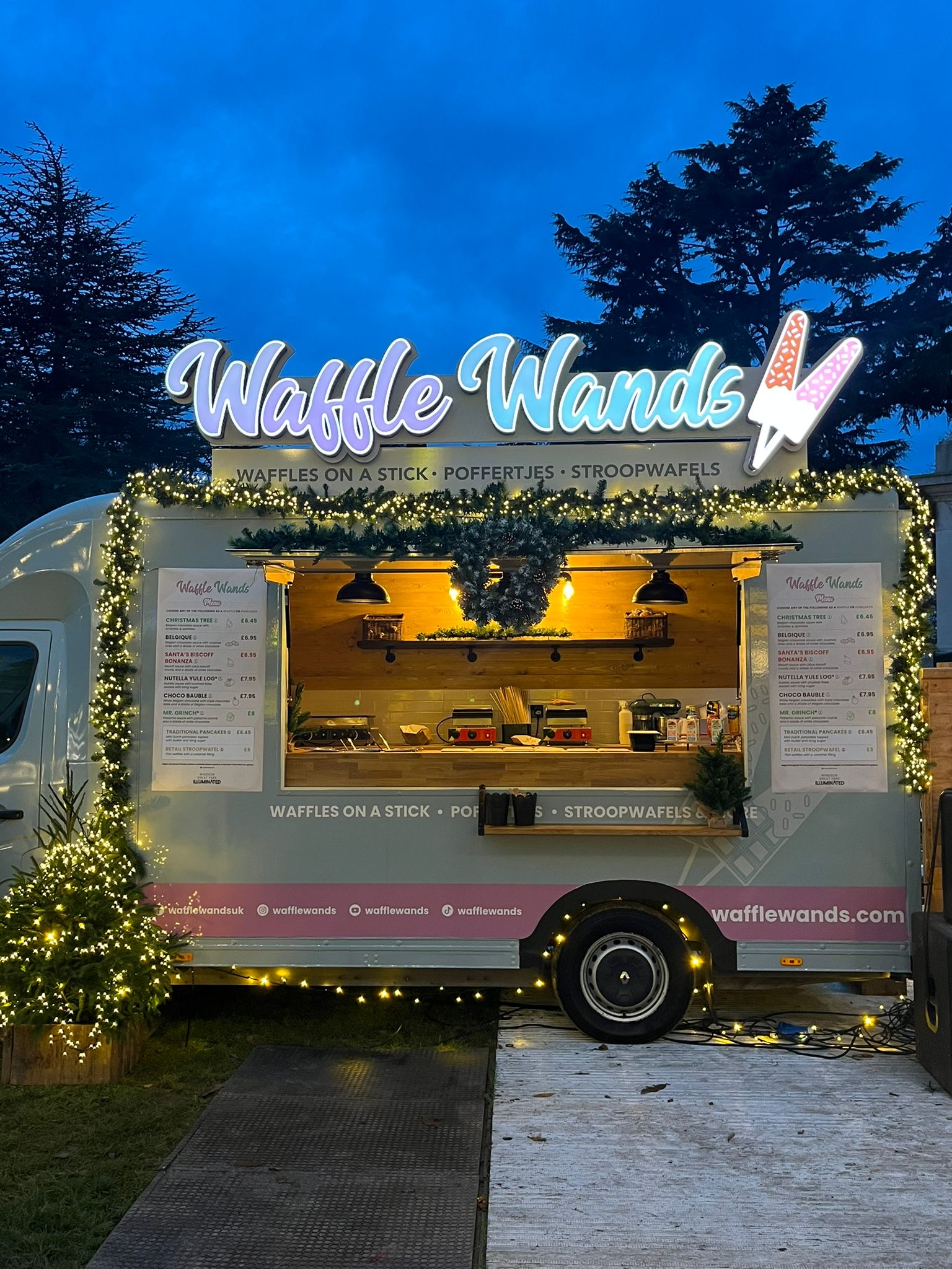 Waffle Wands food truck glowing with fairy lights in St Albans at dusk, serving waffles on a stick from a pastel trailer beneath an illuminated sign.